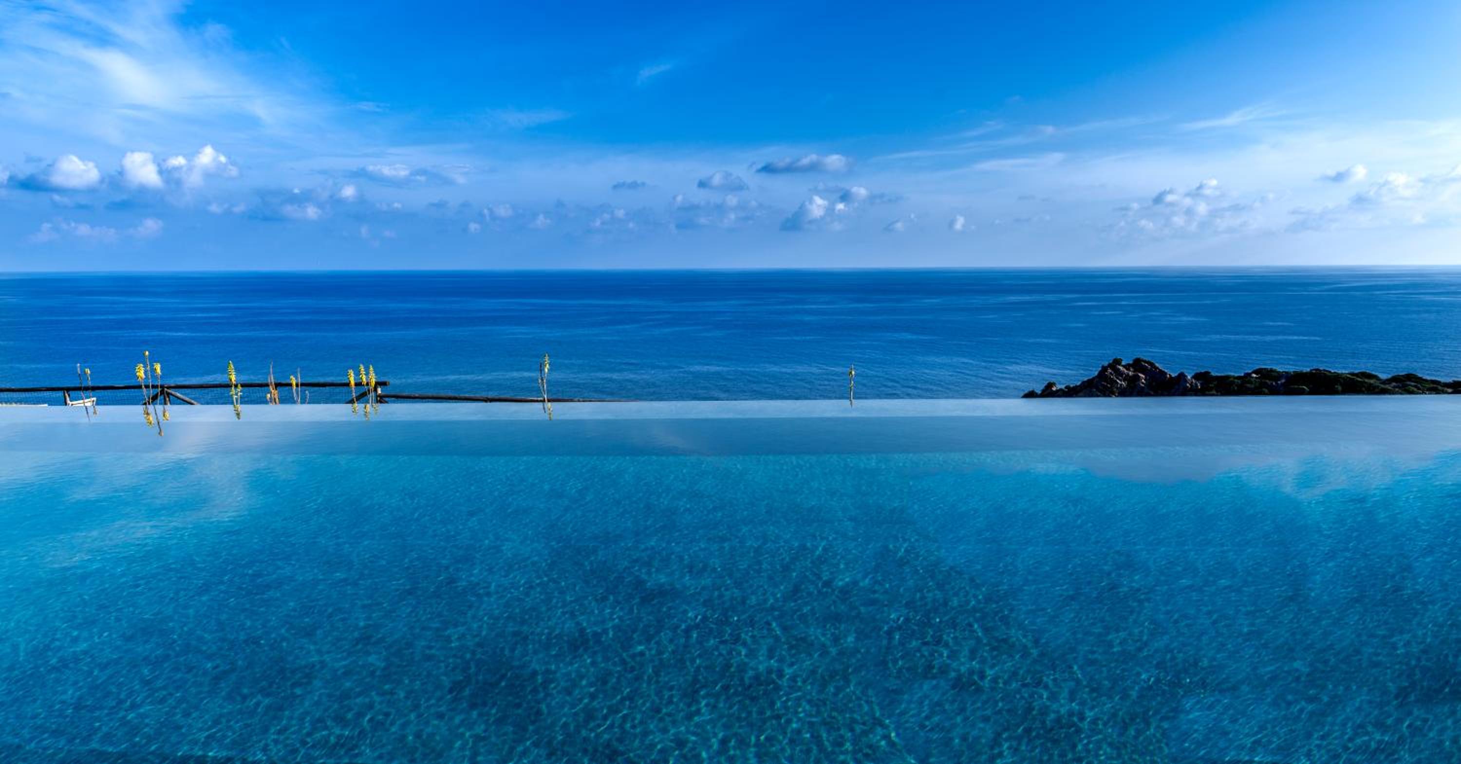 Crystal-clear infinity pool waters mirror the endless azure horizon, where golden posts stand sentinel against dramatic coastal rocks and billowing clouds.