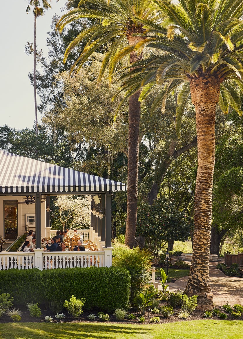 Striped awnings shade al fresco diners beneath towering palms, where California sunshine filters through swaying fronds onto manicured hedgerows and terra cotta walkways.