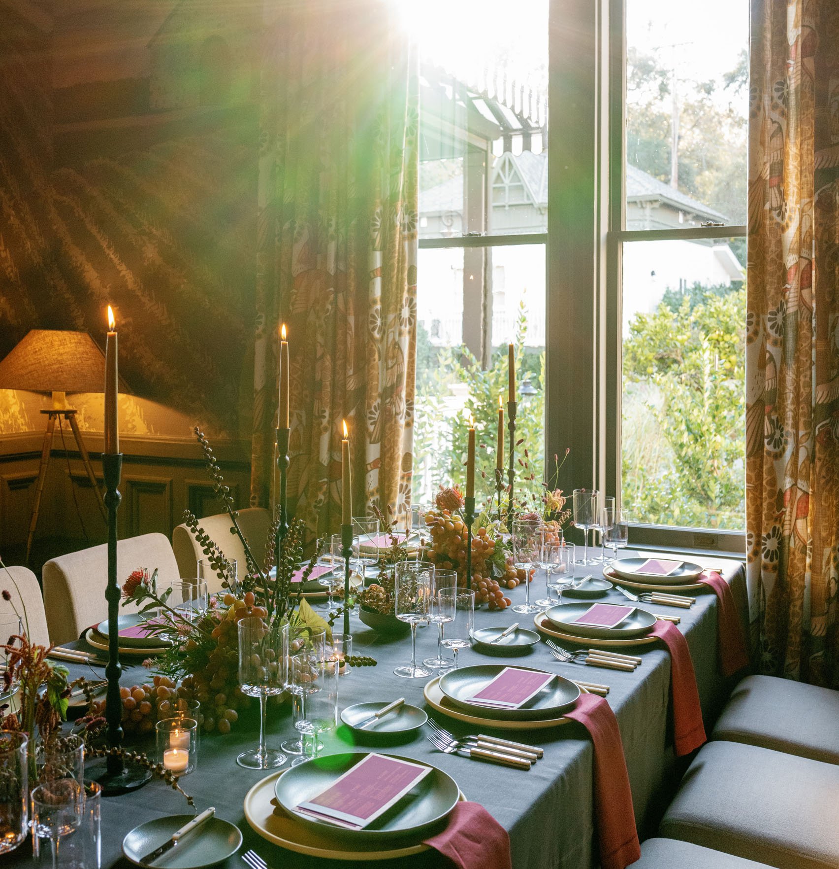 Flickering candlelight dances across an intimate dining table where pink napkins and autumn blooms create warmth against patterned curtains and golden afternoon light.