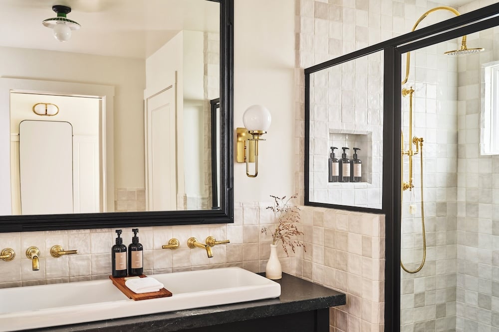 Brass fixtures gleam against handmade zellige tiles in this spa-like retreat — globe sconces, a generous rainfall shower, and curated amenities signal genuine attention to craft.