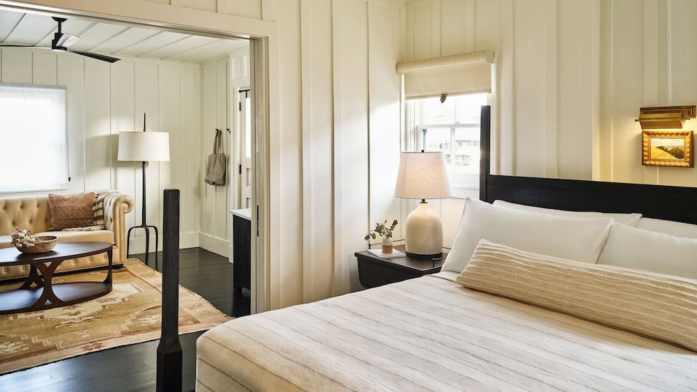 Crisp white shiplap walls and a dark four-poster frame set a quietly elegant tone, where warm lamplight spills across layered linens into a cozy sitting room beyond.