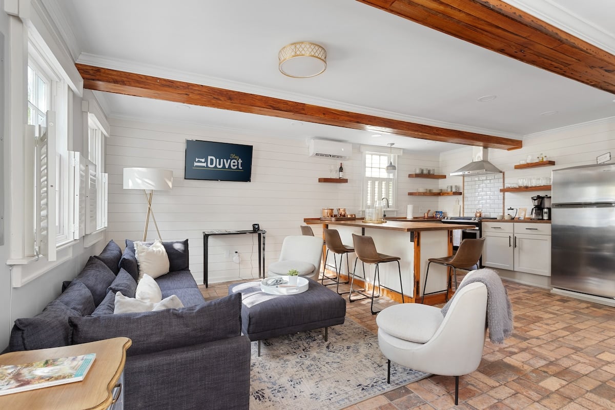 **CAPTION:** Nestled in the heart of Charleston, "Property" showcases a cozy blend of rustic charm and modern elegance. The open-concept space features exposed wooden beams and crisp white shiplap walls, exuding a welcoming and airy atmosphere. Stylish furnishings, including a plush sectional sofa and sleek bar stools, offer a perfect blend of comfort and sophistication. With warm earth tones and natural light, this property provides an inviting escape for discerning travelers seeking both relaxation and style.