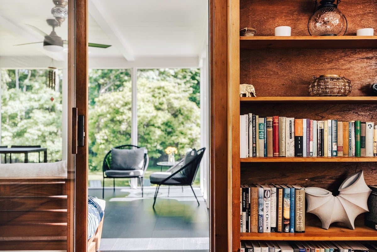 **CAPTION:** Discover the sophisticated charm of "Property" in Woodstock, US, where mid-century modern architecture meets serene nature. The interior boasts warm wooden tones, a well-curated bookshelf, and expanses of glass that invite natural light and lush greenery inside. The stylish yet cozy seating area offers a perfect retreat for discerning travelers seeking tranquility. This luxurious sanctuary promises a harmonious blend of indoor comfort and the beautiful outdoors, ideal for relaxation and inspiration.