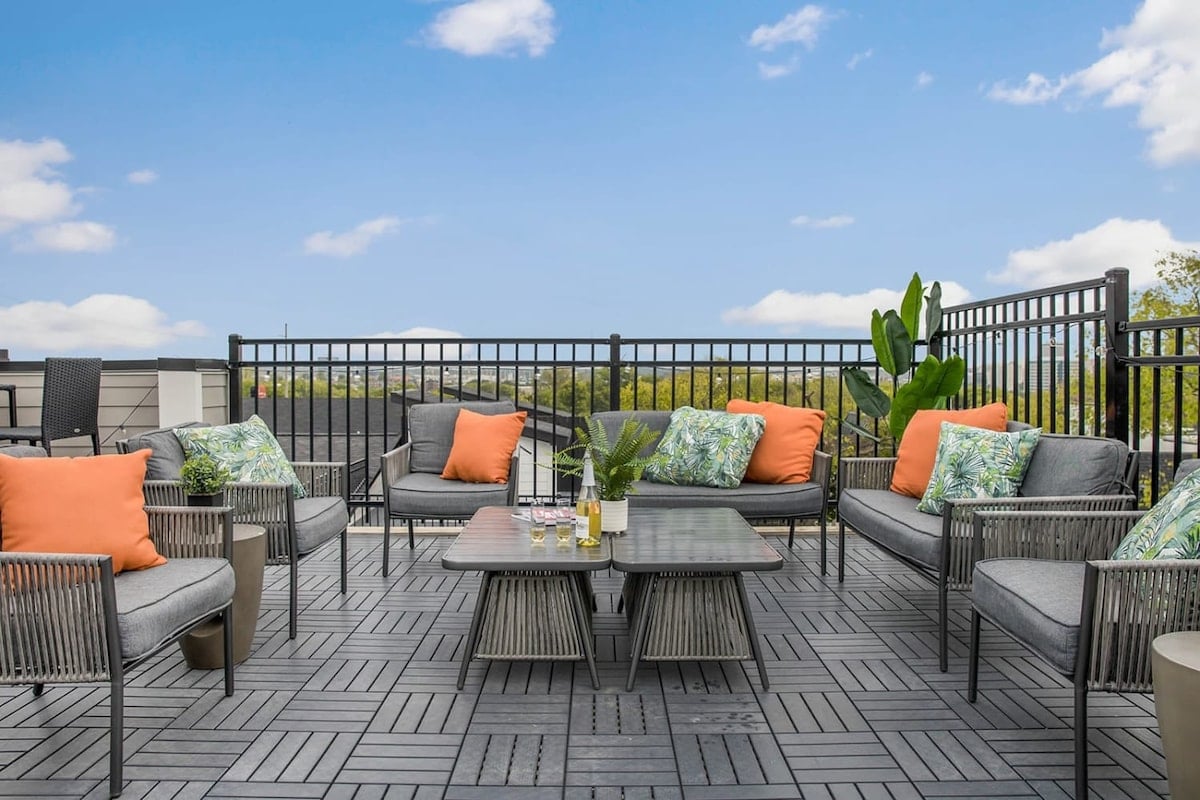 **CAPTION:** Experience a contemporary urban retreat at "Property" in Nashville, US, where modern design meets outdoor comfort. The stylish rooftop patio offers inviting gray wicker seating accented with vibrant orange cushions and lush green plants, providing a serene escape amidst the cityscape. The sleek design and calming color palette create an inviting atmosphere perfect for leisurely gatherings and picturesque views. Ideal for travelers seeking a blend of luxury and relaxation in a vibrant city setting.