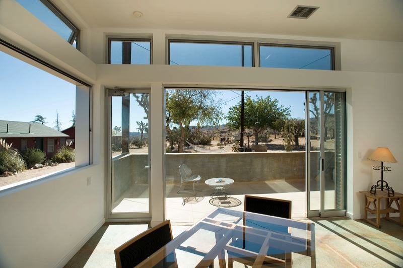CAPTION: 
"Property in Pioneertown showcases a modern architectural style with expansive glass windows offering seamless indoor-outdoor living. The bright and airy atmosphere is complemented by a natural desert landscape visible through sliding doors. Earthy tones and minimalist furnishings enhance the tranquil ambiance, perfect for travelers seeking a serene retreat. With a sunlit terrace and unobstructed views, this luxurious haven invites guests to experience a blend of comfort and nature-inspired elegance."