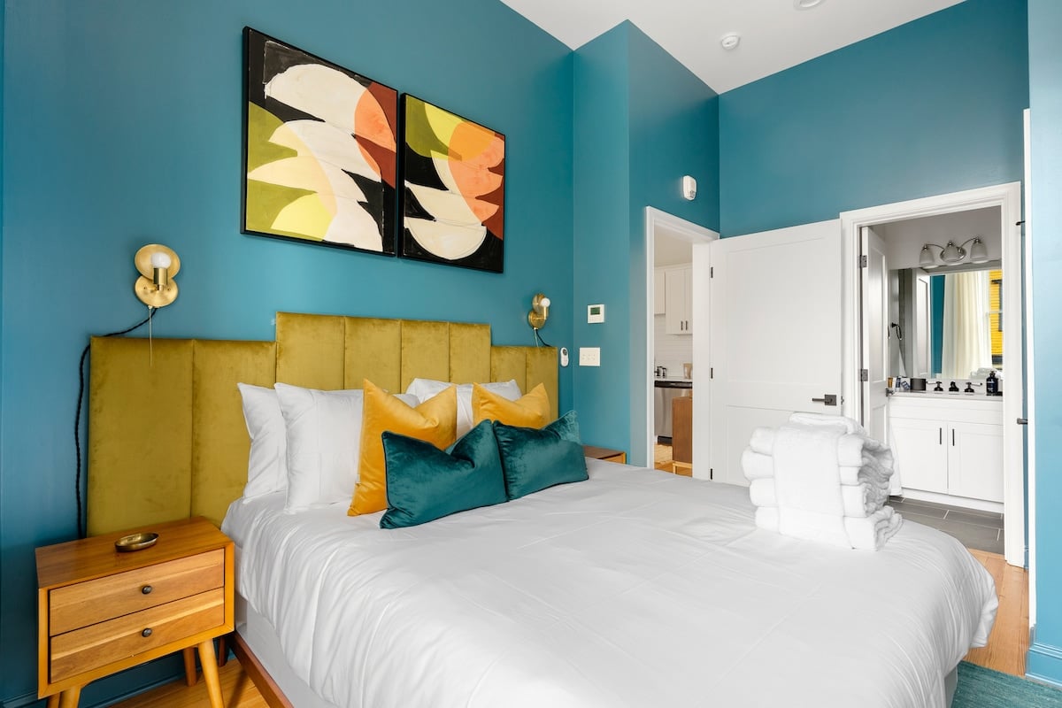 CAPTION:
"Property, a luxury travel destination in Charleston, showcases modern elegance with mid-century accents. The guest room features a striking teal blue palette, complemented by vibrant abstract art and plush velvet headboard, creating a serene yet sophisticated atmosphere. Warm, golden lighting and minimalist wooden furniture enhance the inviting ambiance, appealing to travelers seeking stylish comfort in a refined setting."