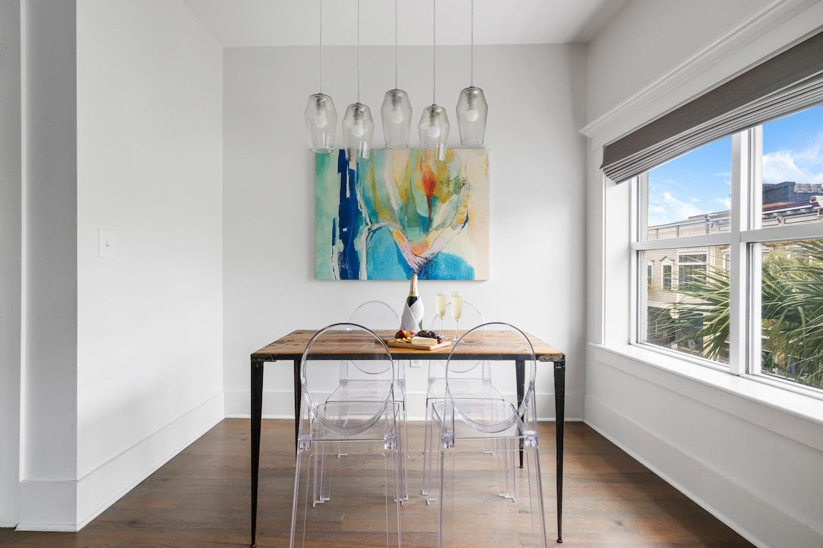 CAPTION: 
The "Property" in Charleston showcases a modern minimalist design, defined by its sleek lines and airy ambiance. The dining area features a rustic wooden table paired with transparent chairs, set against pristine white walls, creating an inviting and serene atmosphere. A splash of color is added by abstract artwork, enhancing the vibrant yet tranquil space. This blend of contemporary and classic elements appeals to travelers seeking a chic and serene luxury escape.