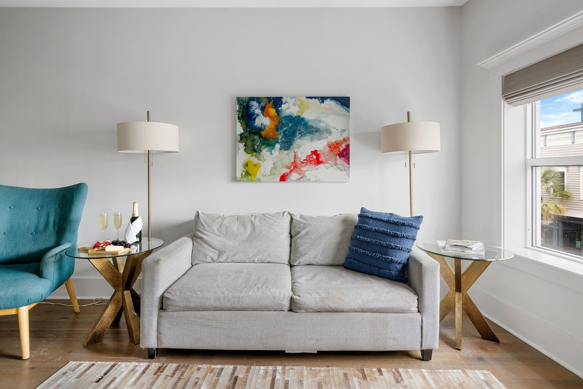 CAPTION: Nestled in the heart of Charleston, the "Property" exudes a modern, minimalist charm with its clean, contemporary design. The serene atmosphere is enhanced by a soft neutral color palette, featuring gray and teal accents. Elegant furnishings, including a chic gray sofa and stylish armchairs, provide a comfortable retreat. The vibrant abstract artwork adds a splash of color, appealing to travelers seeking sophistication and comfort in a luxury setting.