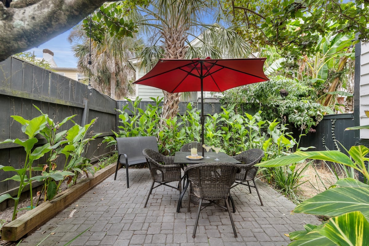 CAPTION: Discover unparalleled luxury at "Property" in Charleston. This inviting outdoor retreat boasts contemporary charm with a cozy, brick-paved patio. Surrounded by lush greenery, the space features a striking red umbrella and comfortable wicker seating, creating a serene oasis for relaxation. The vibrant green tones of the foliage contrast beautifully with the muted gray tones of the patio, crafting a calm yet sophisticated ambiance ideal for travelers seeking a peaceful escape.