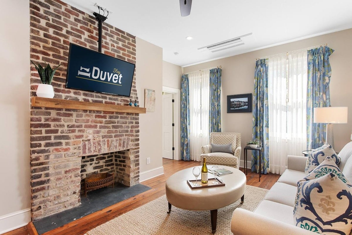 CAPTION: Nestled in the heart of Charleston, "Property" exudes Southern charm with its classic brick fireplace and cozy, inviting atmosphere. The space harmoniously blends traditional design with contemporary elegance, featuring plush furnishings and vibrant accents. Warm earthy tones are complemented by bursts of color from decorative elements. Ideal for travelers seeking a luxurious yet homey retreat, "Property" offers a quintessential Charleston experience that delights with its timeless beauty and modern comforts.