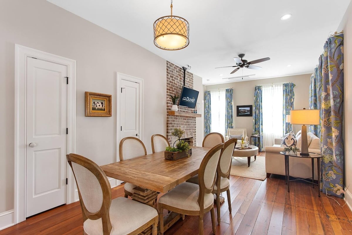 CAPTION: The Property in Charleston showcases a harmonious blend of historical charm and modern comfort. The warm, earthy tones of the wooden flooring complement the exposed brick fireplace, creating an inviting atmosphere. Classic wooden furniture juxtaposes elegantly with contemporary lighting, while patterned curtains add a vibrant touch. This stylish space marries rustic elegance with a cozy vibe, perfect for travelers seeking a luxurious yet home-like experience in the heart of Charleston.