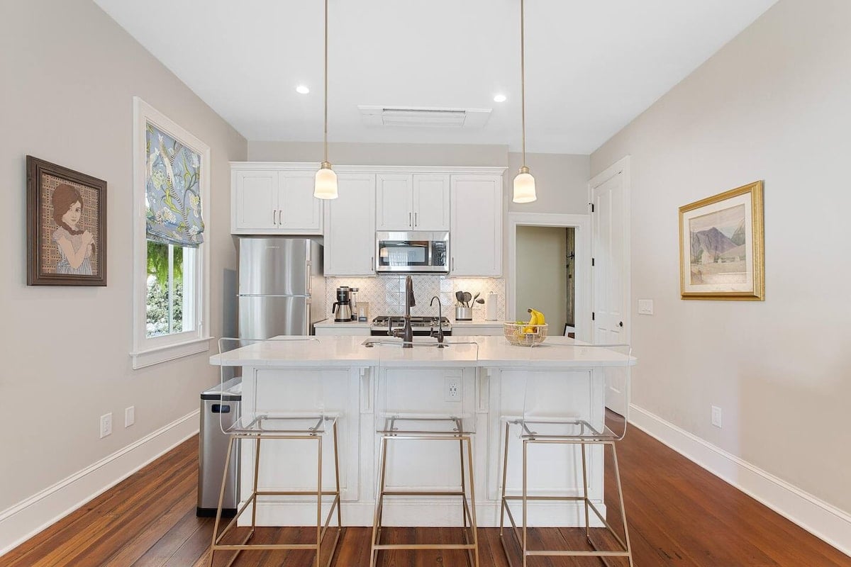 CAPTION: This luxurious Charleston property showcases a modern yet cozy atmosphere, perfect for upscale travelers. The open-concept kitchen is adorned with sleek stainless steel appliances and a crisp white color palette, contrasted beautifully by rich, warm hardwood floors. Minimalist pendant lighting enhances the chic, contemporary vibe, making it an inviting space. Ideal for those seeking a refined yet comfortable experience in elegant Southern surroundings.
