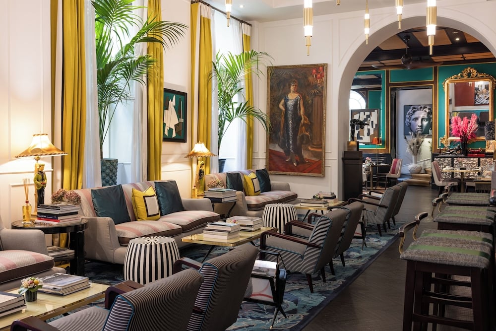 Golden curtains frame intimate seating vignettes beneath classical portraiture, where striped ottomans and jewel-toned furnishings create a sophisticated Roman salon atmosphere.