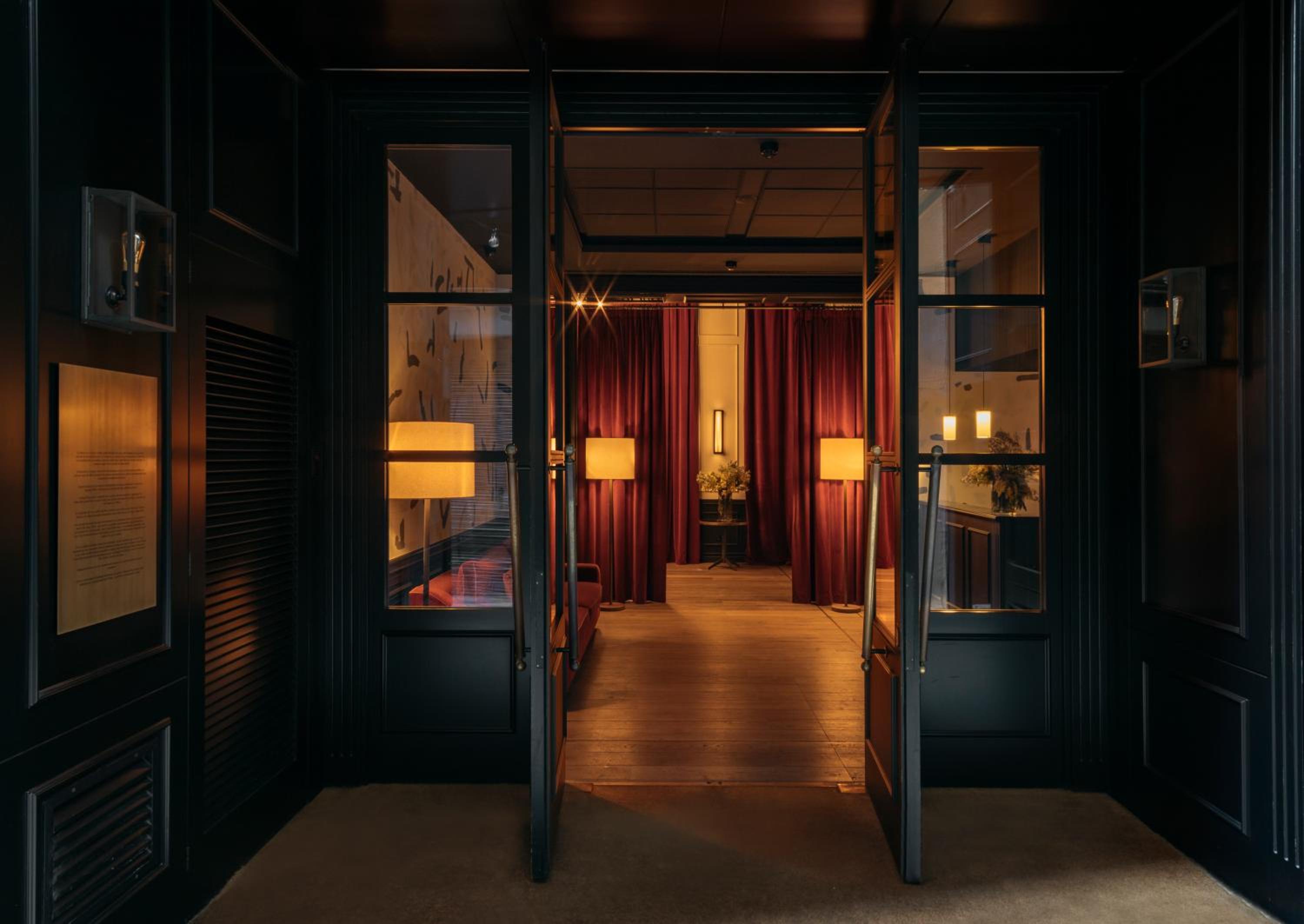Dramatic red velvet curtains frame the intimate entrance, where warm amber lighting and sleek black frames create an inviting passage between sophisticated spaces.