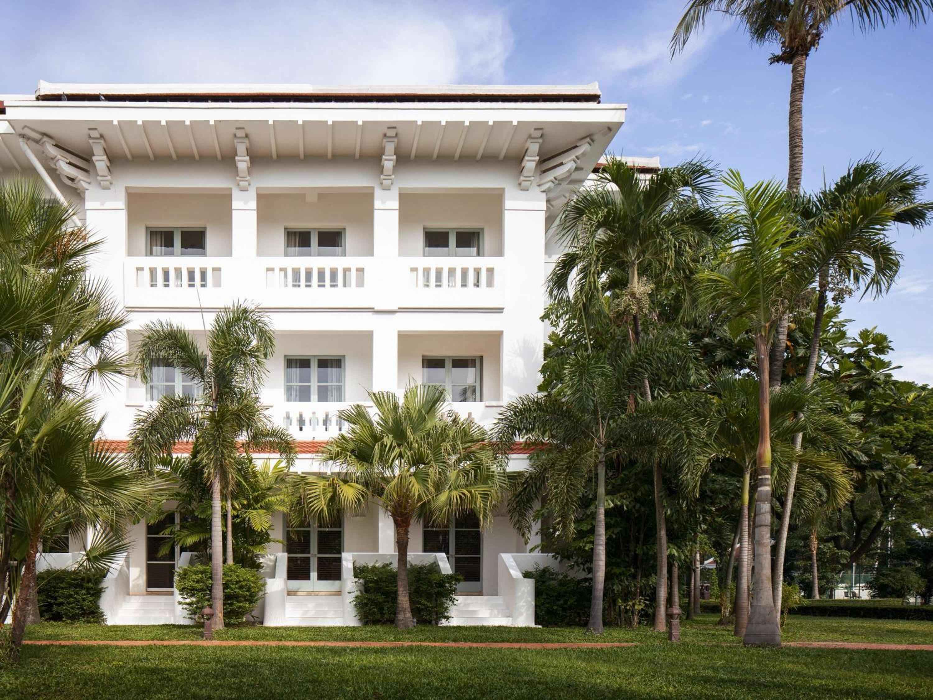 Colonial-era elegance radiates from pristine white facades framed by swaying coconut palms, where manicured lawns and traditional Khmer architecture create an oasis of refined tranquility.