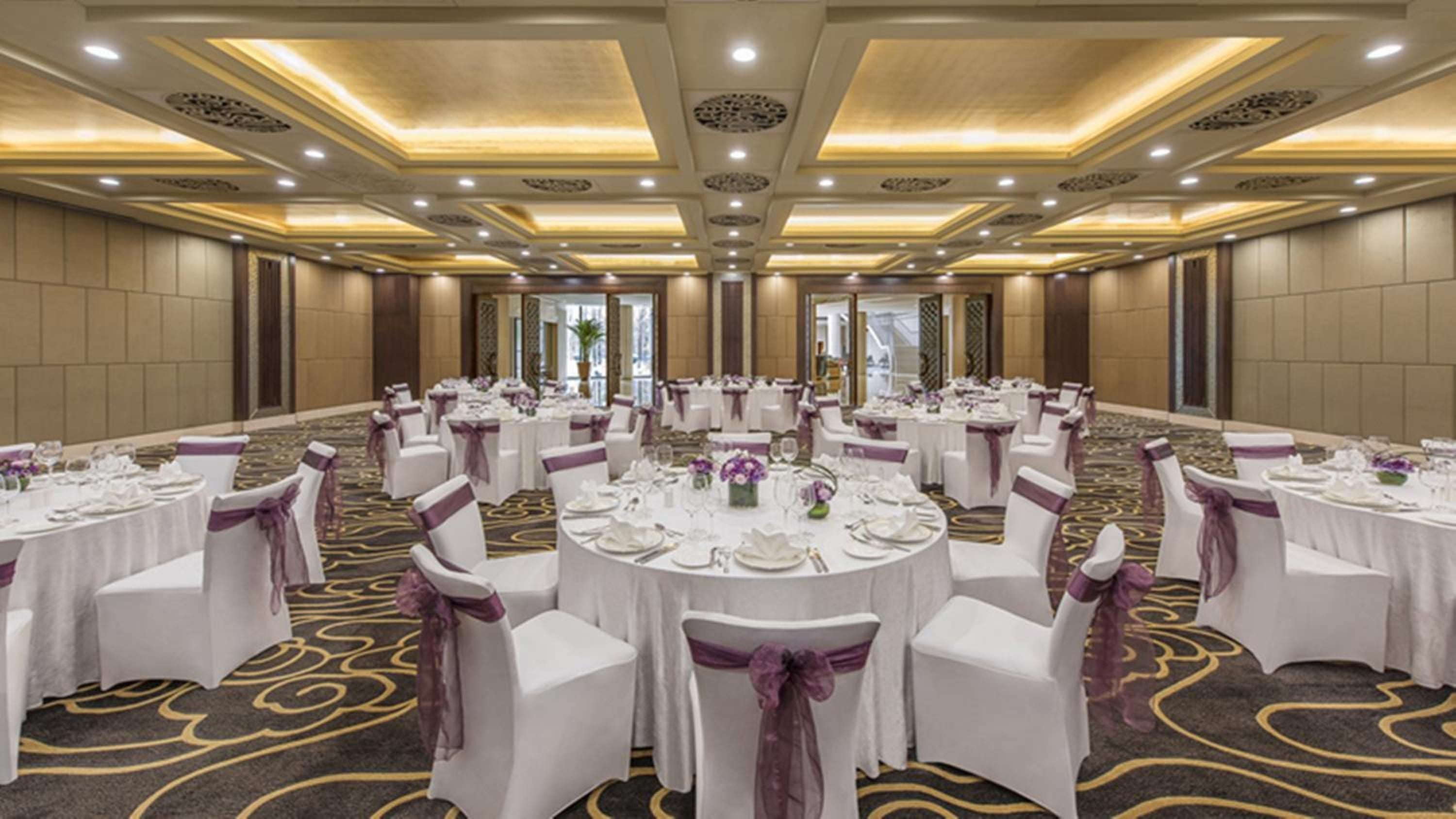 Coffered ceilings with warm amber lighting frame an elegant ballroom where purple sashes add sophistication to crisp white linens and gleaming place settings.