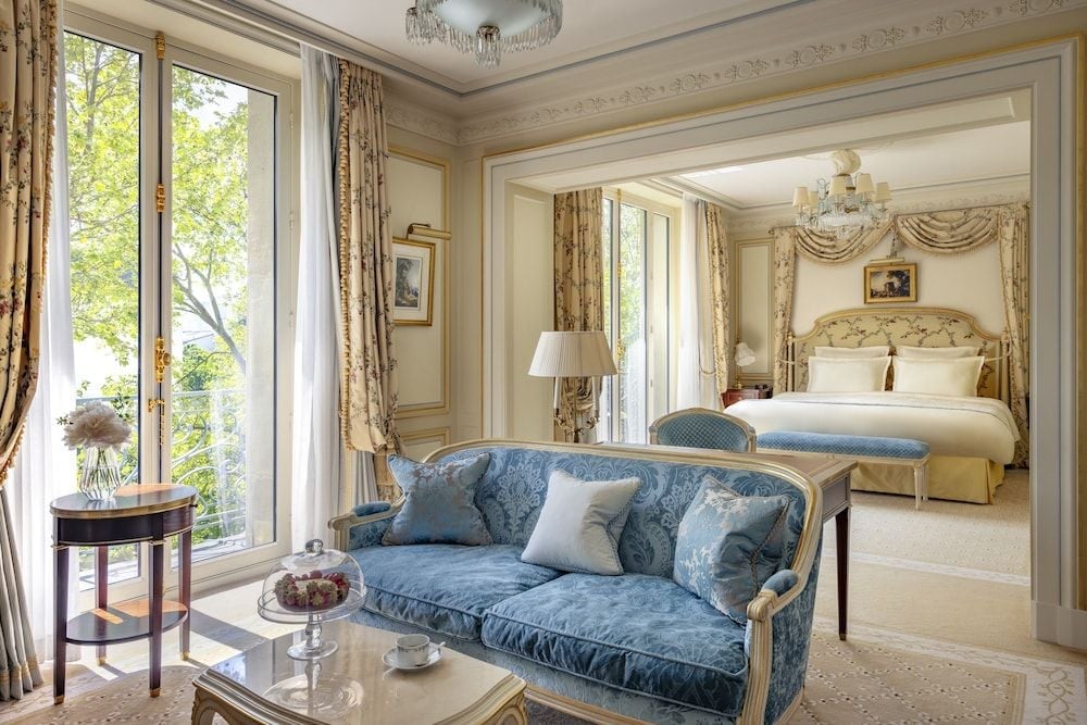 Gilded moldings frame sapphire silk furnishings in this palatial suite, where crystal chandeliers cast gentle light across cream carpets and garden views beyond.