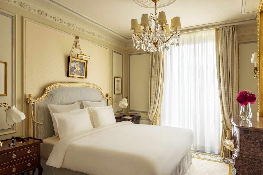 Gilded moldings frame sumptuous cream linens while crystal chandeliers cast warm light across this palatial guest room, where Parisian elegance meets timeless luxury.