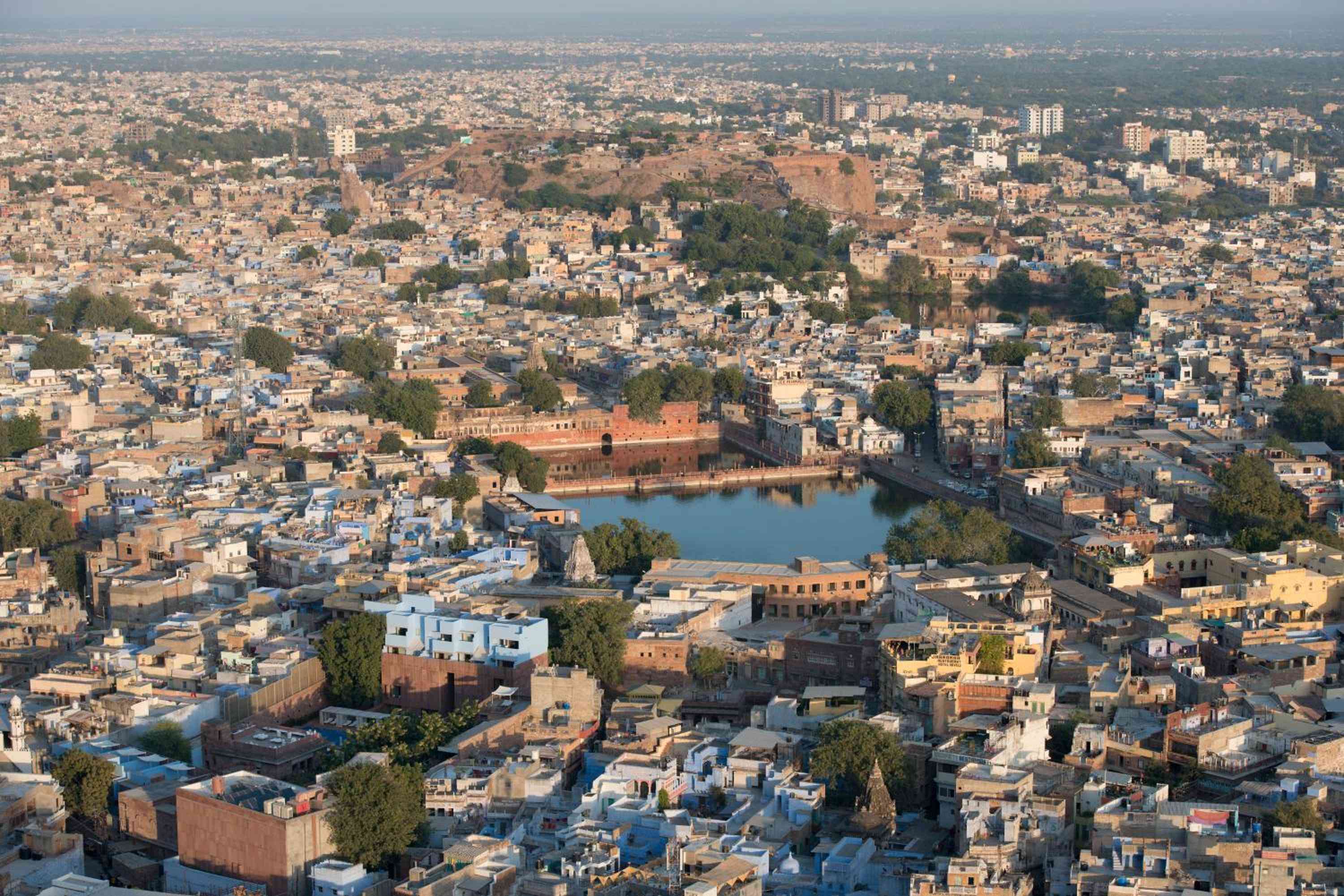Ancient Jodhpur unfolds in sandstone splendor from above, its historic fort walls embracing a tranquil lake while the Blue City's maze of rooftops stretches endlessly toward distant hills.