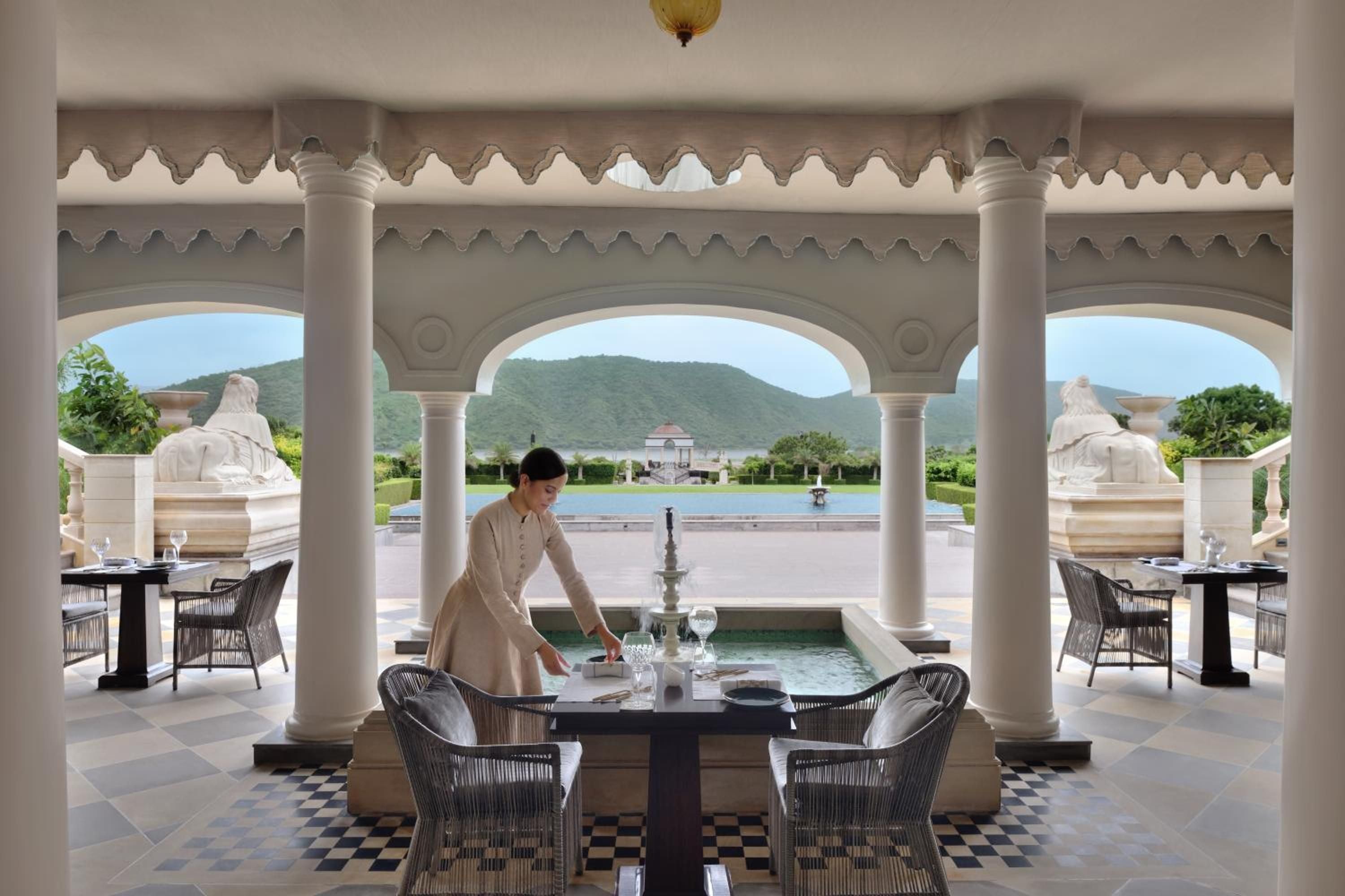 Beneath ornate Mughal arches, a server sets tables overlooking tranquil pools and distant Aravalli hills, where marble elephants guard an palatial dining pavilion.
