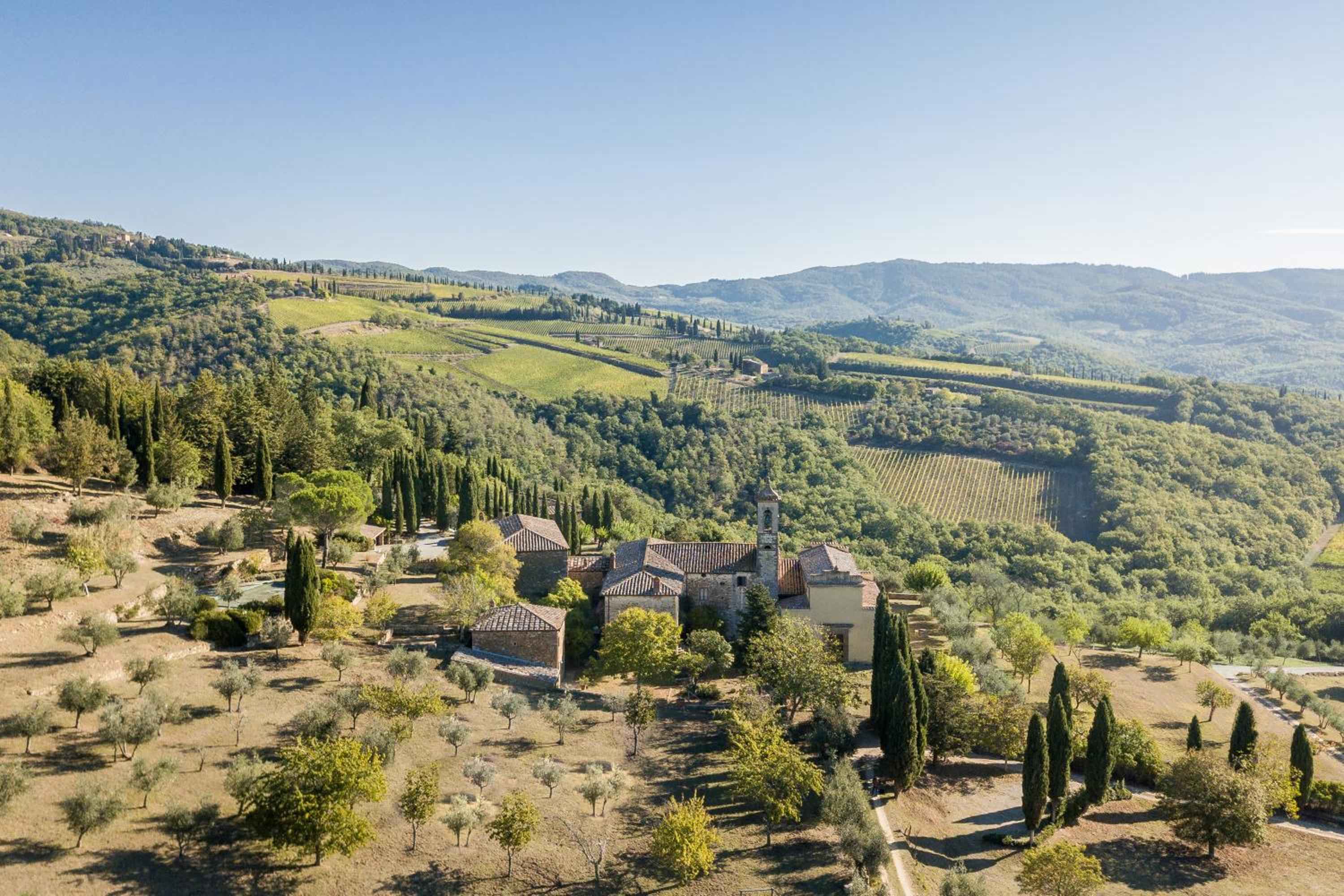 Stone buildings crowned with terracotta tiles nestle among cypress groves and olive trees, while vineyard-striped hills roll toward distant Tuscan peaks under golden light.
