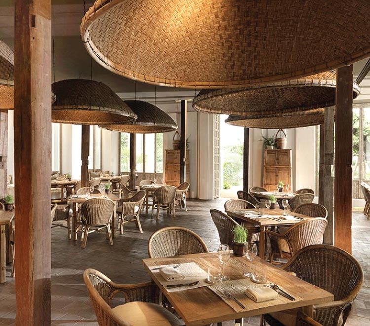 Oversized woven lampshades cast warm shadows across the open-air dining room, where natural materials and floor-to-ceiling windows create an inviting fusion of comfort and craftsmanship.