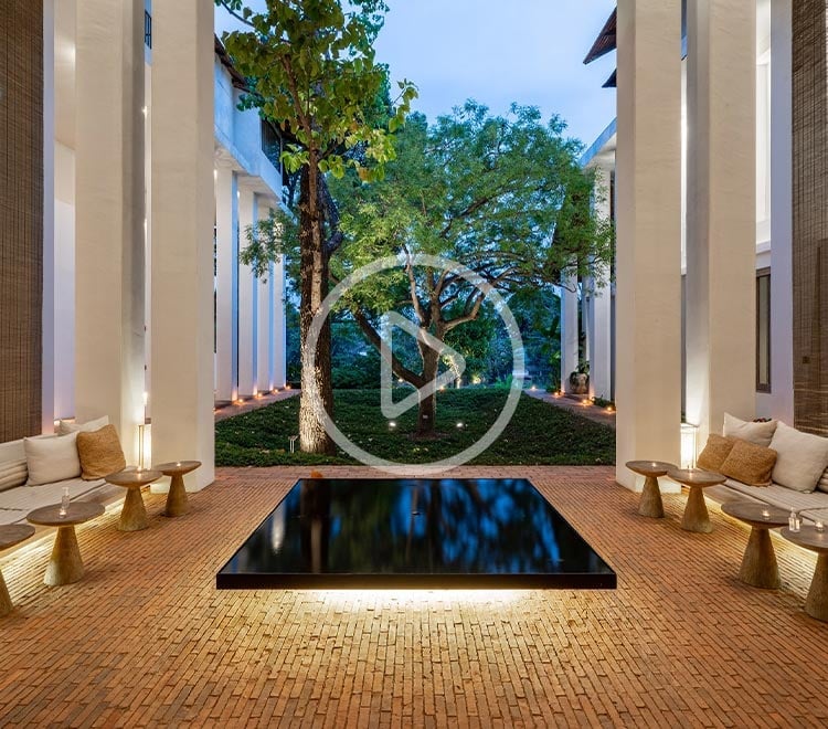 Illuminated columns frame a serene reflecting pool where ancient trees cast shadows on honey-toned brick, creating an intimate courtyard sanctuary within Raya Heritage's contemporary Lanna architecture.
