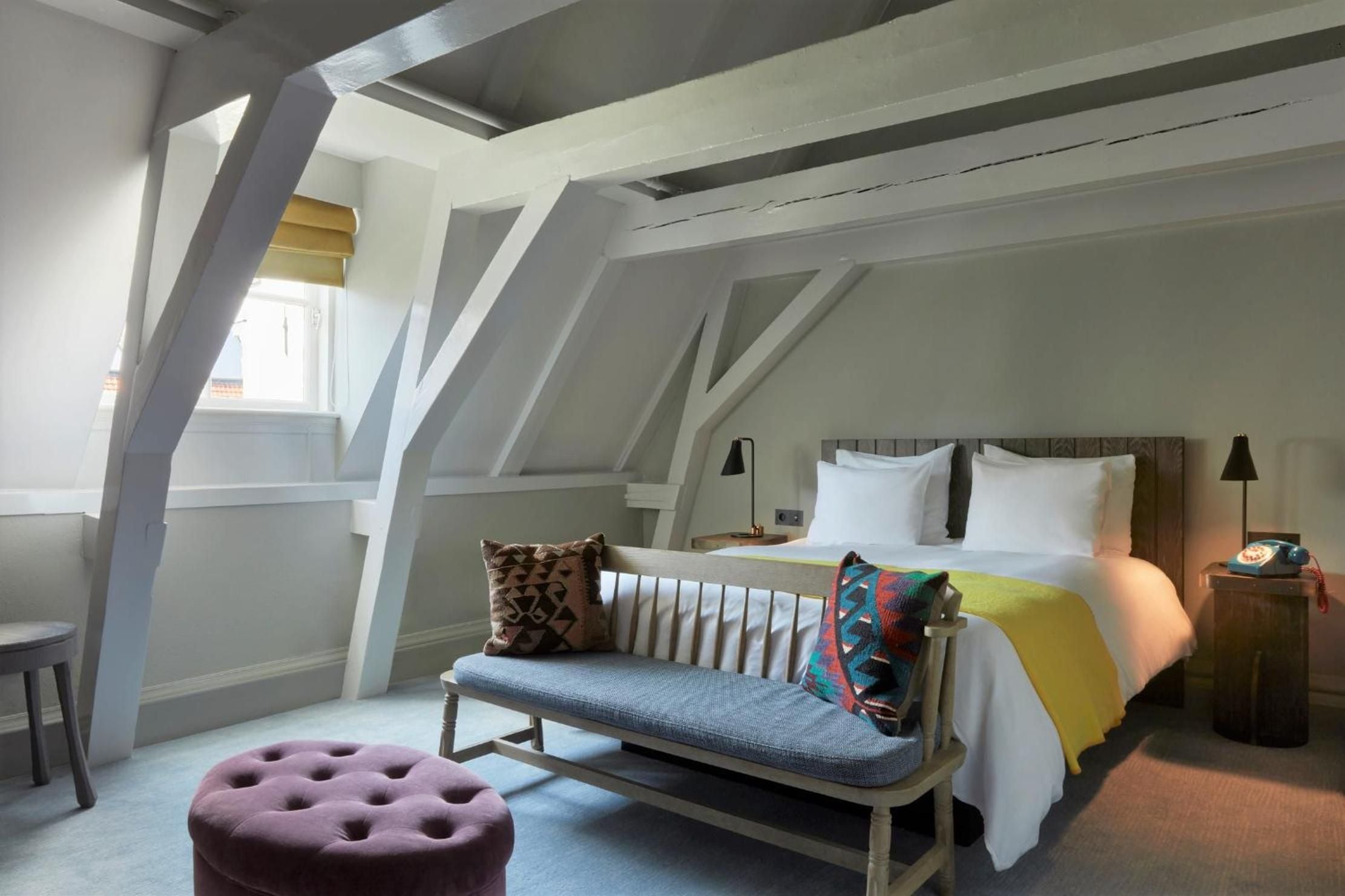 Exposed timber beams frame this charming attic suite, where mustard yellow accents and jewel-toned textiles create intimate warmth beneath Amsterdam's historic rooflines.