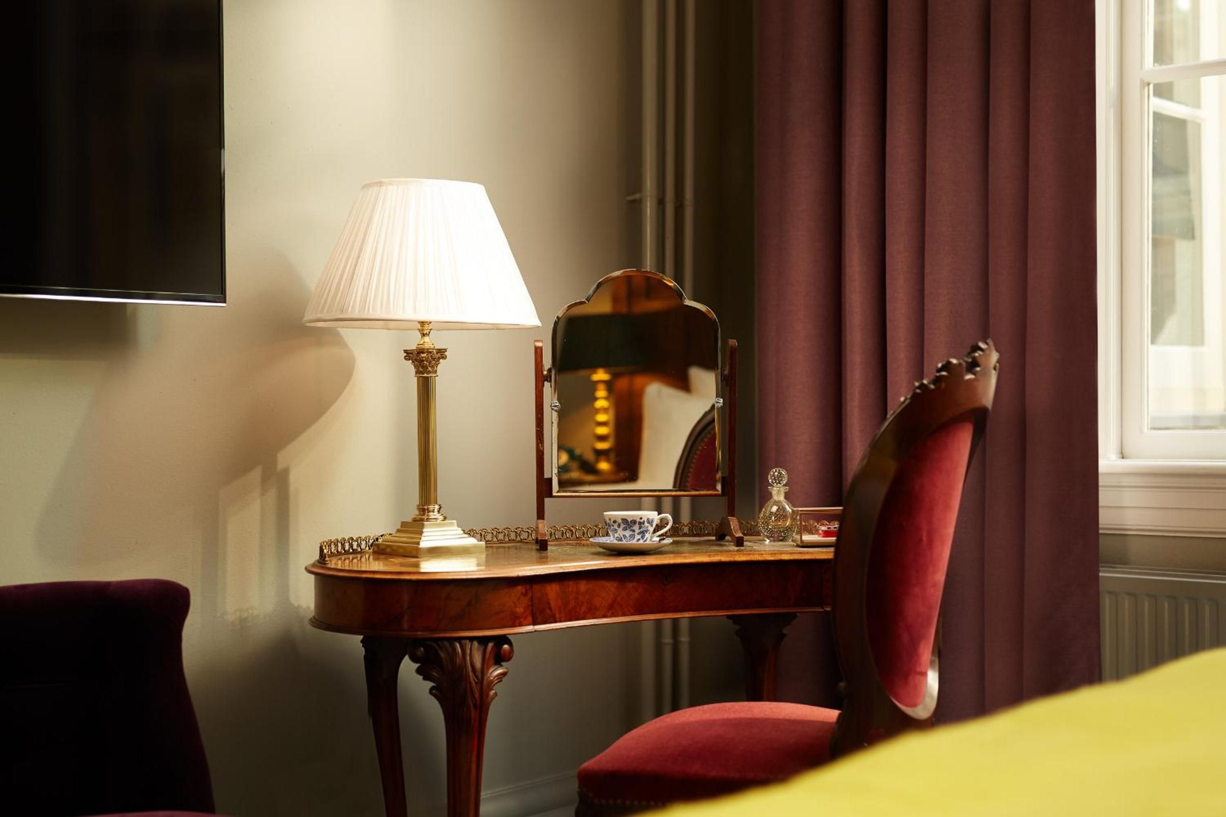 Golden afternoon light illuminates a mahogany writing desk adorned with delicate porcelain and brass accents, where burgundy curtains frame moments of quiet luxury.