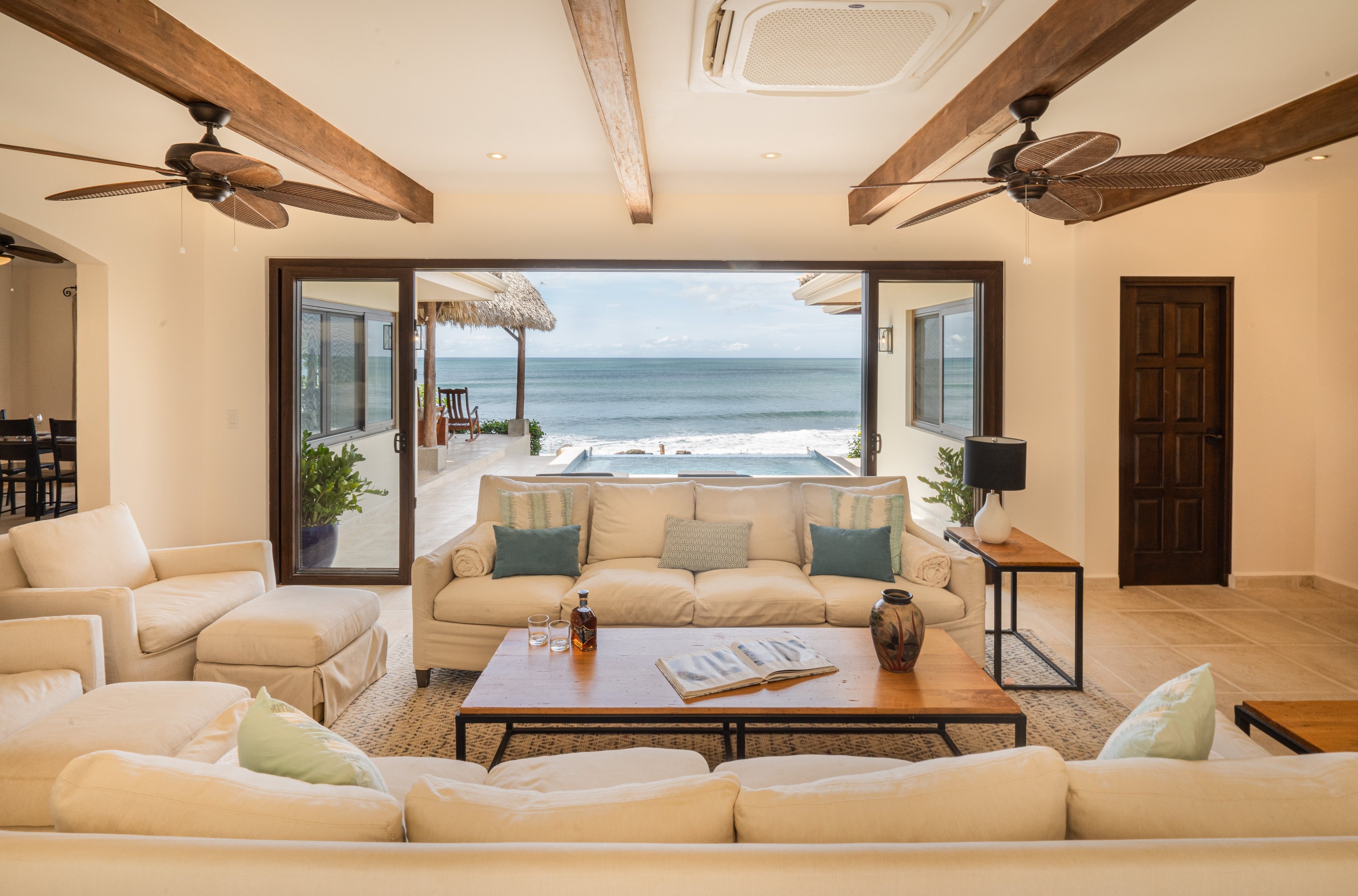 Exposed wood beams frame an open-concept living space where cream sofas invite lingering over morning coffee while Pacific waves crash just beyond the infinity pool.