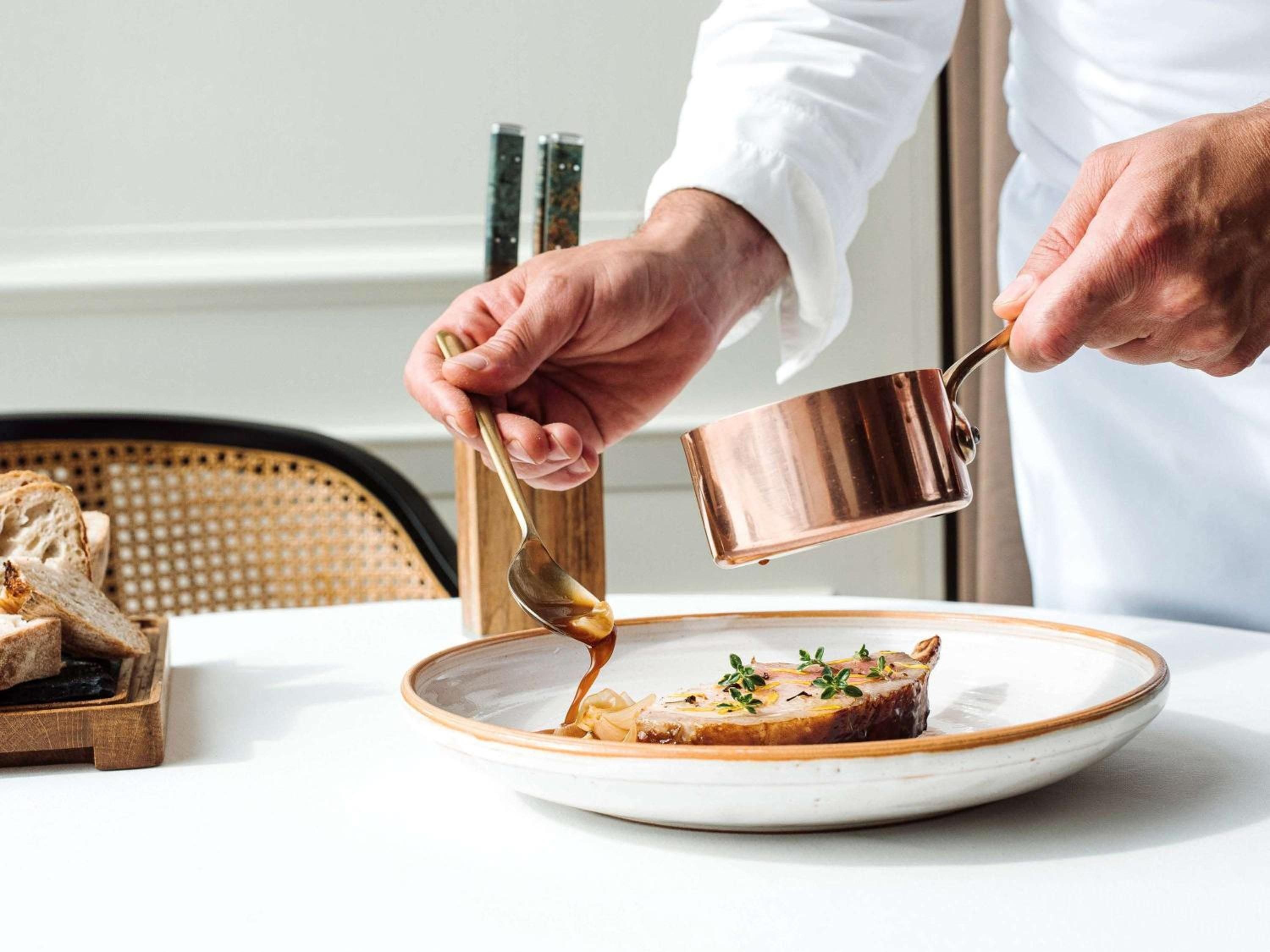 Copper gleams as skilled hands drizzle golden sauce over perfectly plated cuisine, where culinary artistry meets refined European dining tradition in Warsaw's most storied hotel.