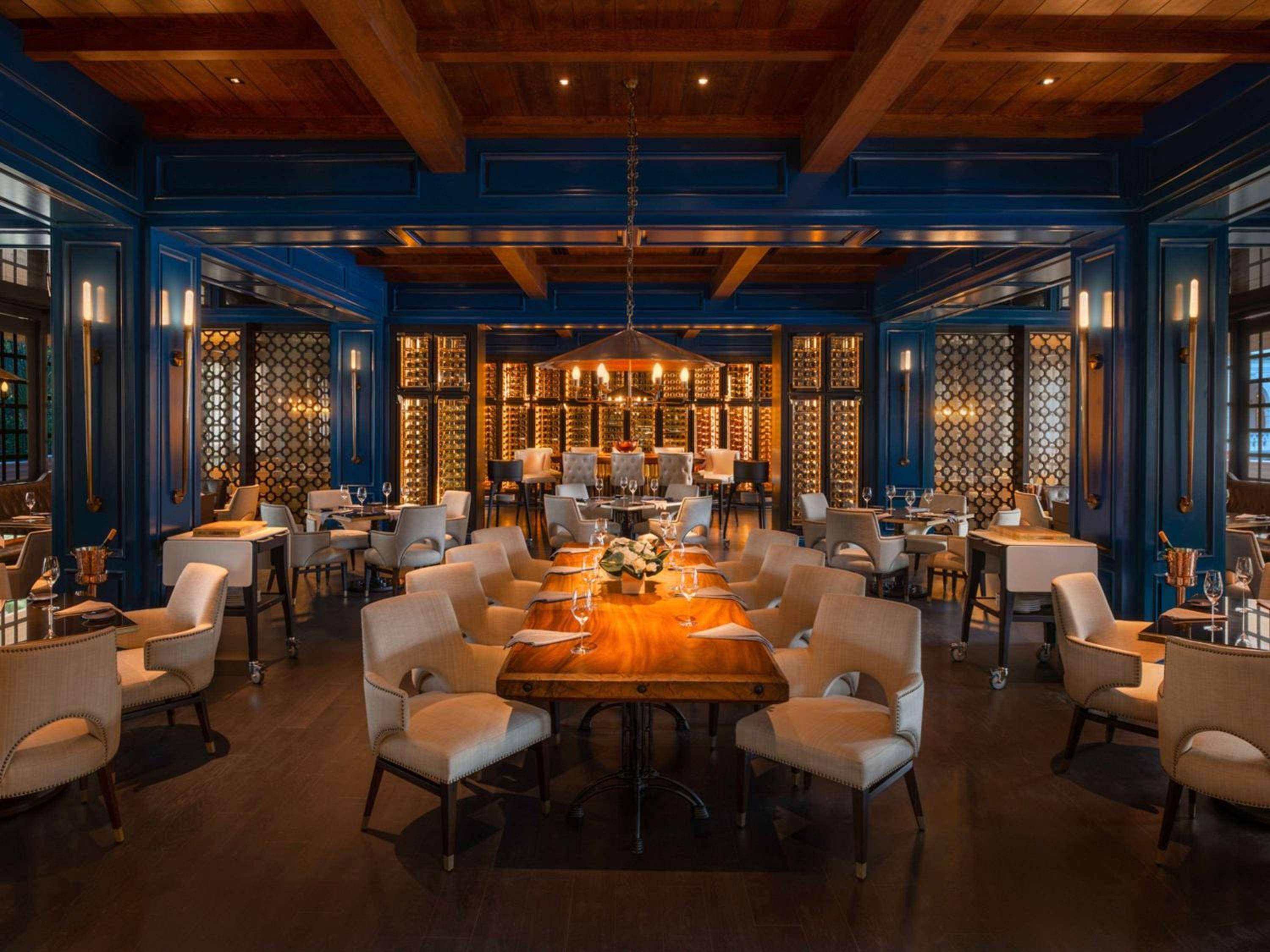 Navy blue paneling and exposed timber beams frame this sophisticated dining room, where live-edge wood tables catch candlelight beneath an ornate wine display wall.