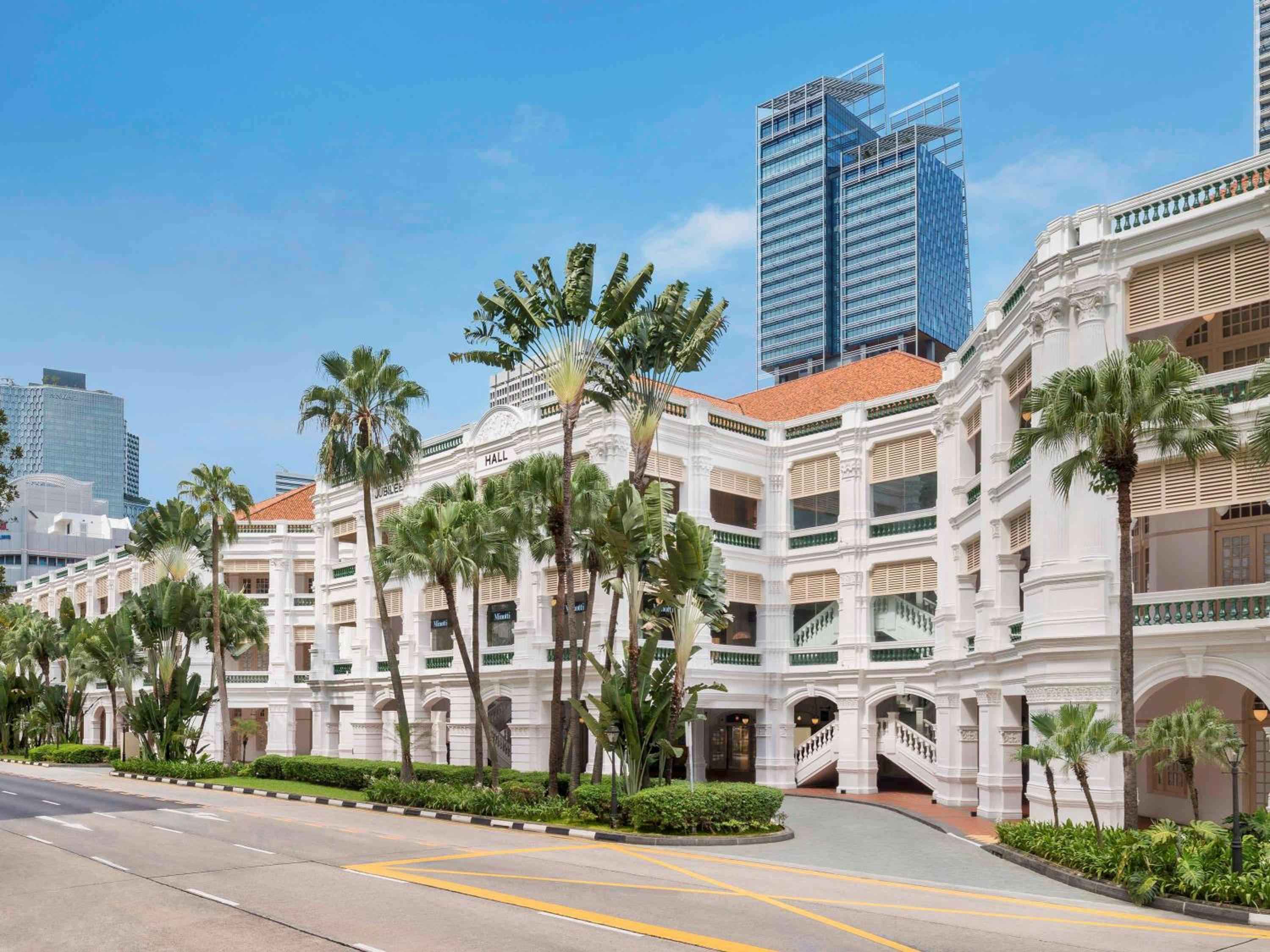 Colonial grandeur meets tropical elegance as palm fronds sway against Raffles' pristine white facade, while Singapore's gleaming towers rise majestically beyond this timeless sanctuary.