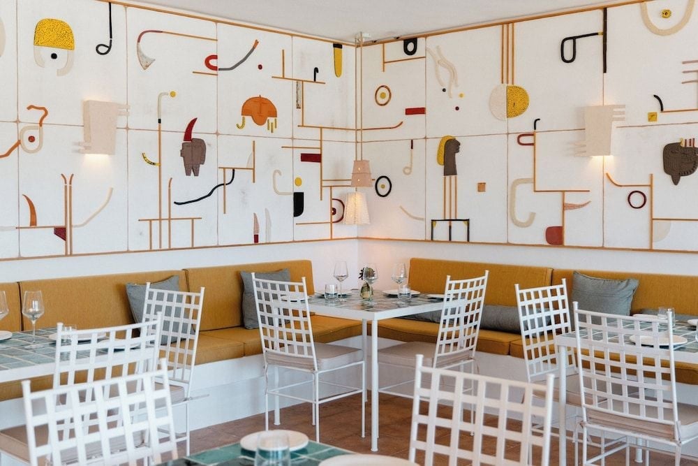 Playful abstract murals dance across white walls while guests settle into mustard banquettes and grid-back chairs, creating an artistic dining atmosphere that feels both sophisticated and whimsical.