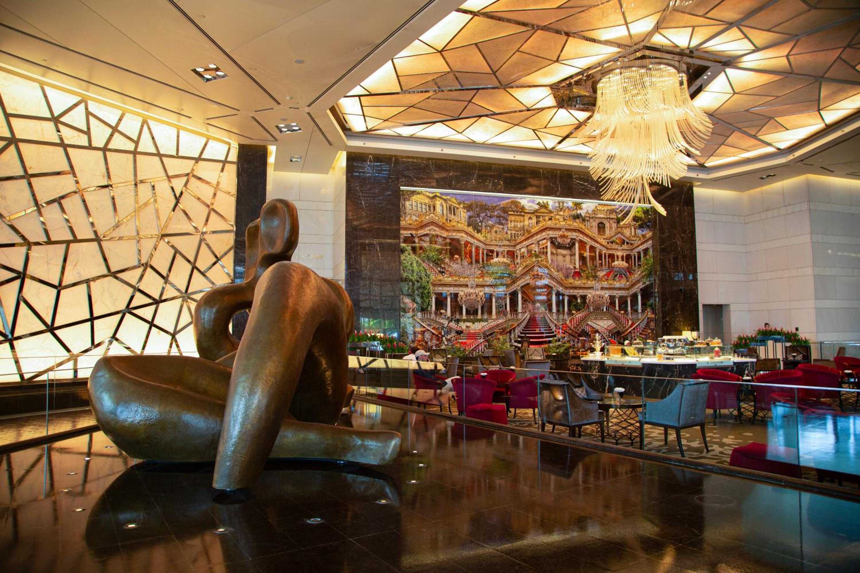 Dramatic geometric skylights bathe the soaring lobby in golden light, where contemporary sculpture meets jewel-toned seating beneath a cascading crystal chandelier.