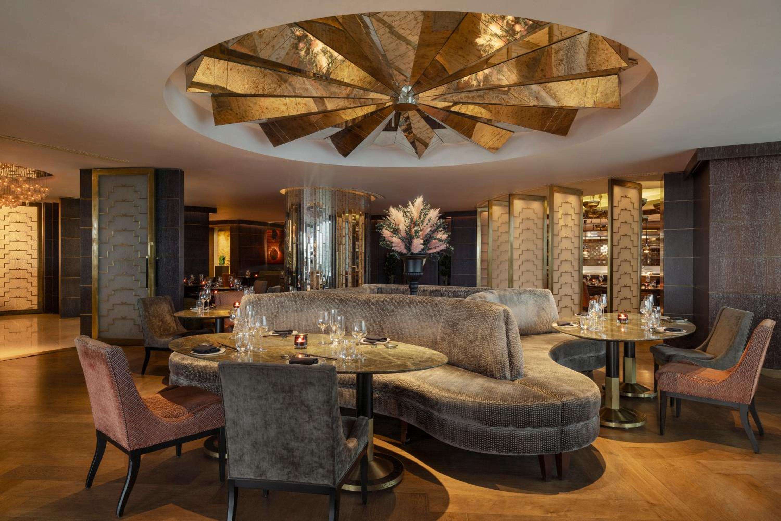 Curved banquettes and warm wood paneling create intimate dining alcoves beneath a striking sunburst ceiling feature, where golden light dances across polished surfaces.