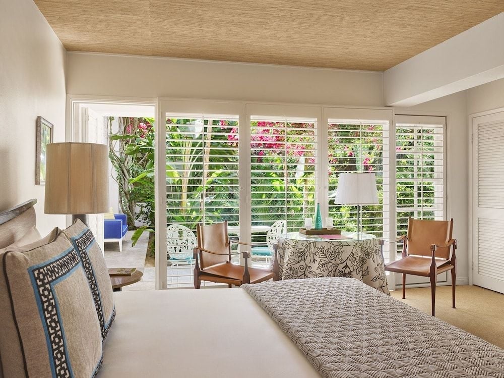Natural light filters through plantation shutters into this serene guest room, where leather chairs frame garden views and a wood-planked ceiling adds Palm Springs warmth.