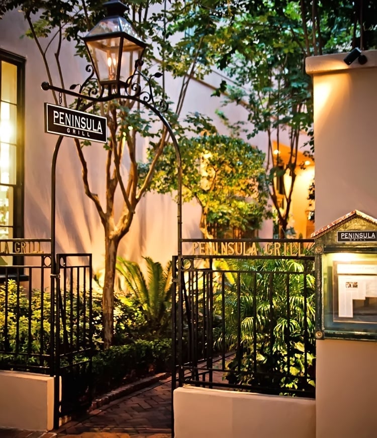 Peninsula Grill's lantern-lit entrance beckons through wrought-iron gates, where moss-draped trees and brick pathways create an intimate Charleston courtyard sanctuary.