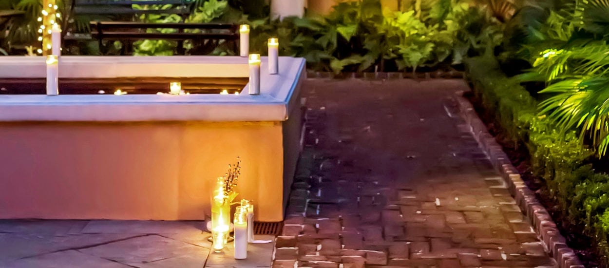 Candles flicker against warm stone walls in the intimate courtyard, where brick pathways wind through lush plantings beneath Charleston's golden evening light.