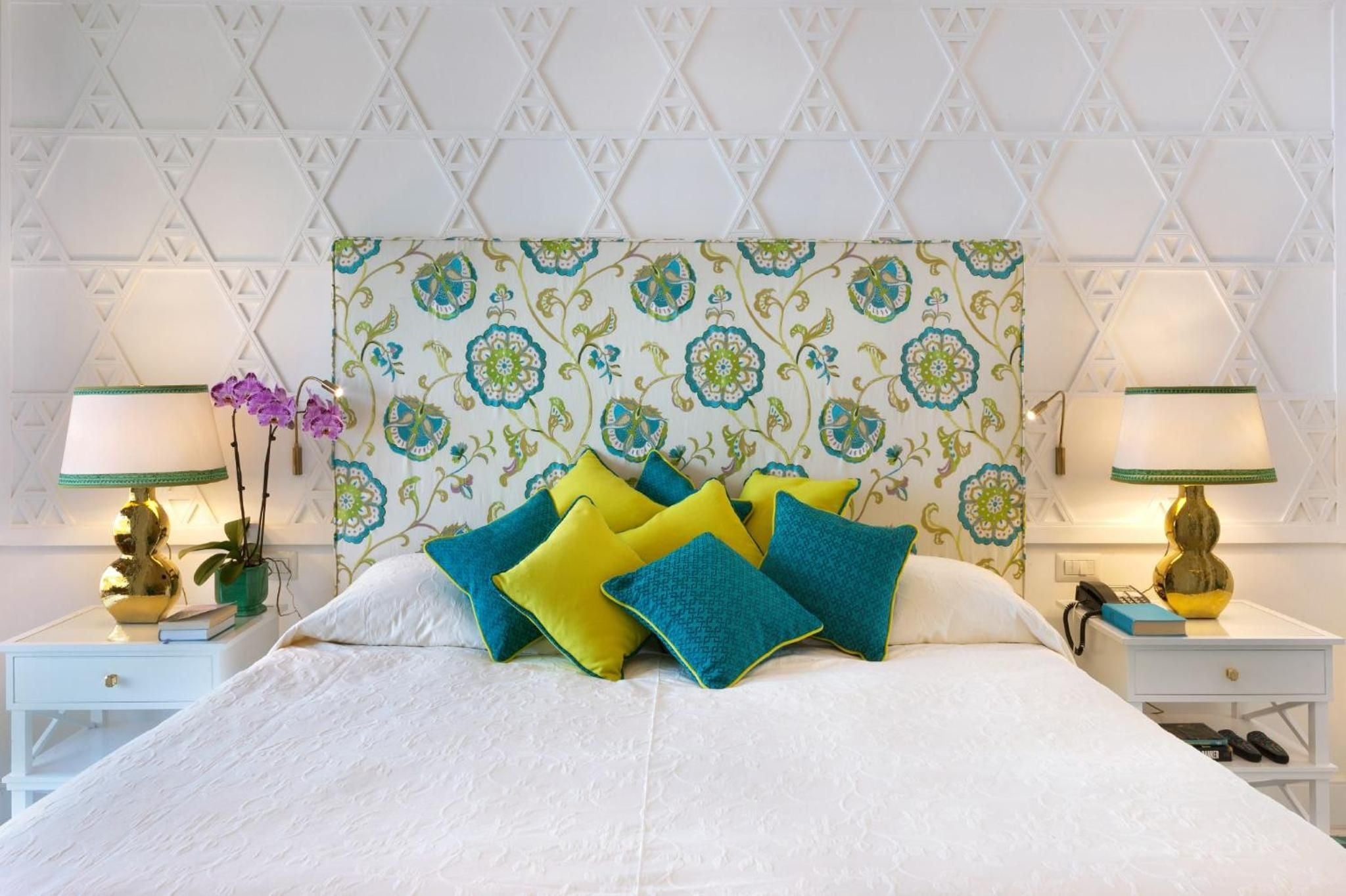 Vibrant botanical headboard fabric meets crisp white linens and jewel-toned pillows, creating an elegant Mediterranean sanctuary with pineapple-base lamps casting warm evening light.