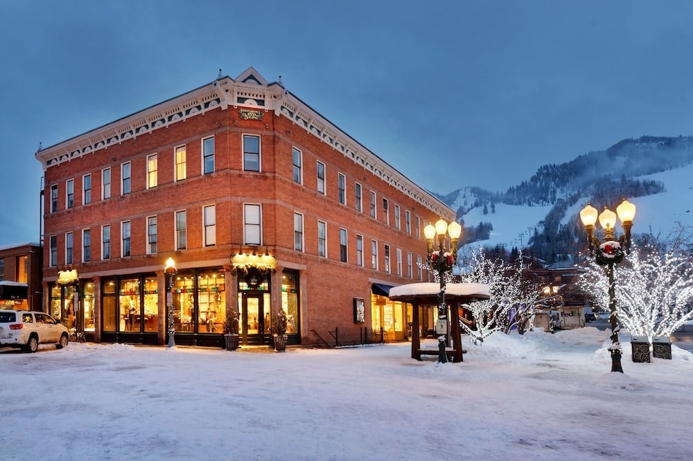 Snow blankets Aspen's historic streets as the iconic brick Residences building glows warmly against twilight skies, with twinkling lights creating magical mountain town ambiance.