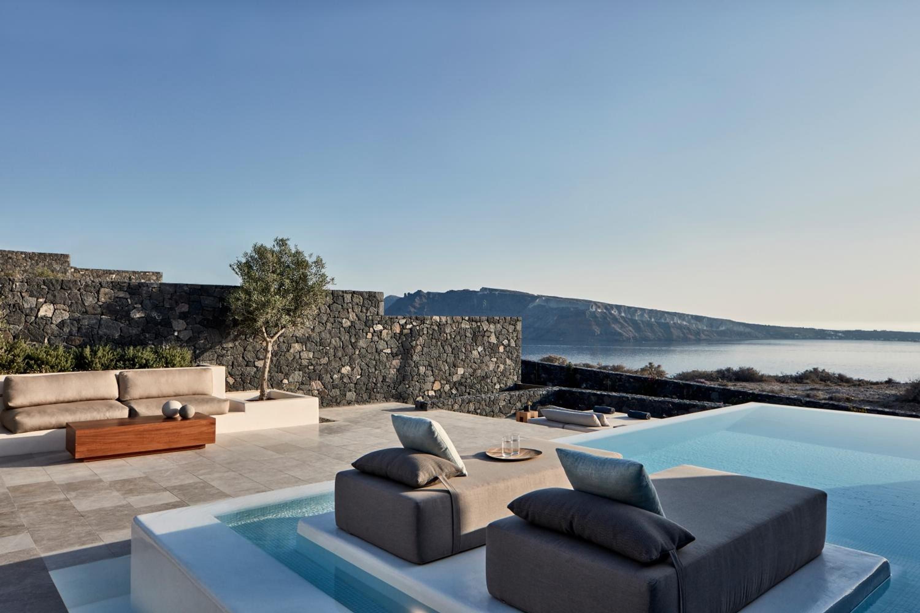 Charcoal loungers float like islands in the infinity pool, where volcanic stone walls frame endless Aegean views and distant caldera cliffs.