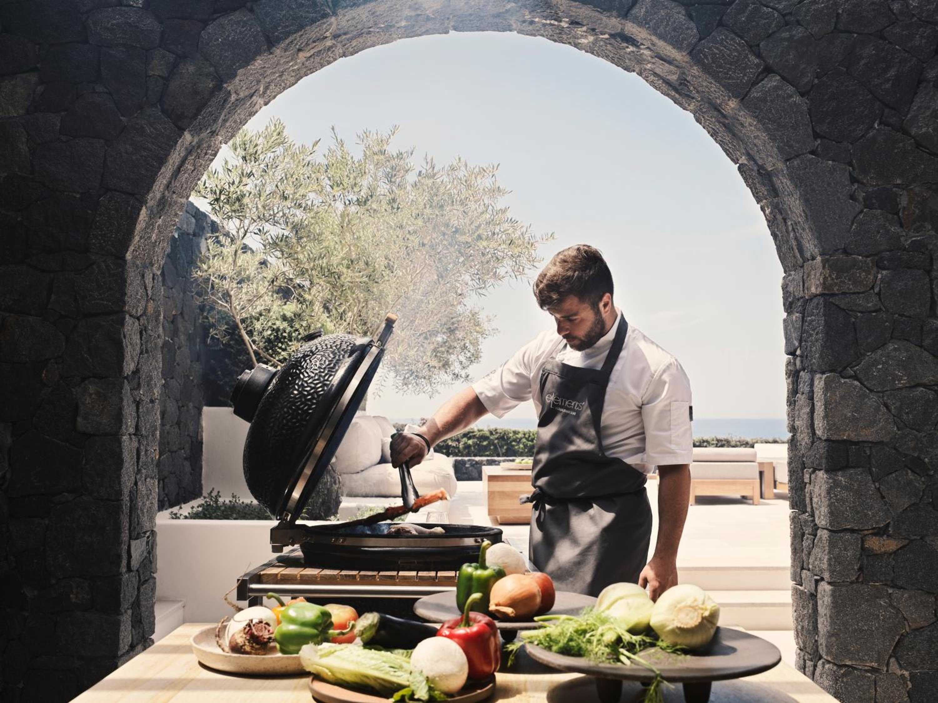 Beneath ancient stone archways, the chef tends to fresh Mediterranean produce at the outdoor grilling station, where Aegean breezes carry the scent of olive oil and herbs.