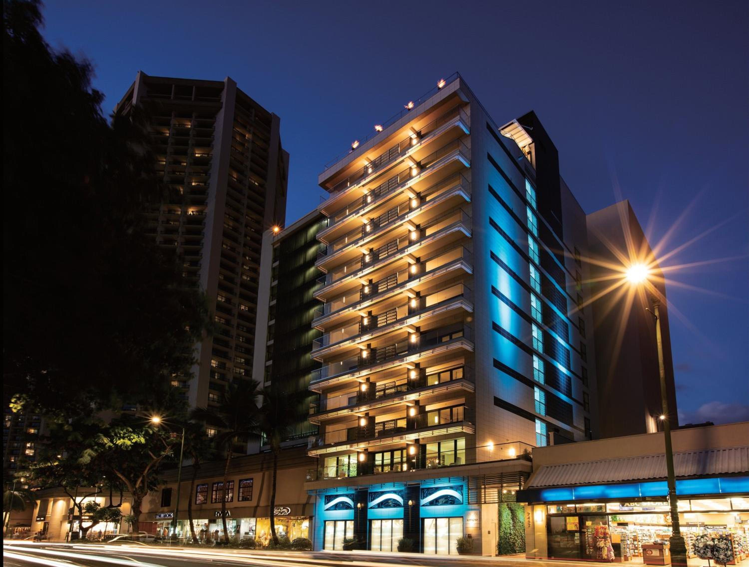 Blue-lit facades and warm balcony lighting create an inviting evening glow along this bustling street, where modern architecture meets vibrant ground-floor retail energy.