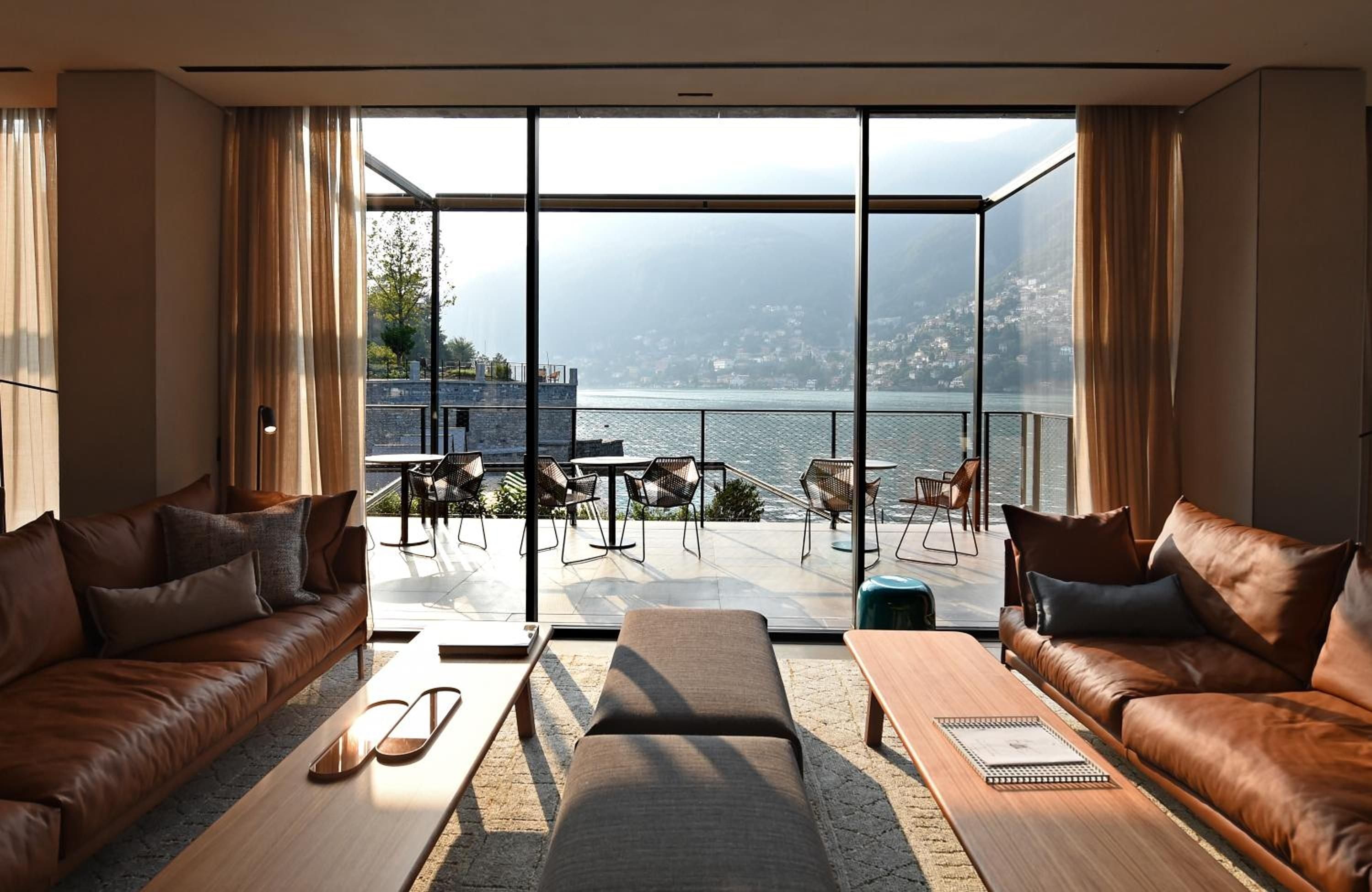 Cognac leather sofas and floor-to-ceiling windows frame Lake Como's dramatic shoreline, where morning light dances across handwoven rugs and warm wood surfaces.
