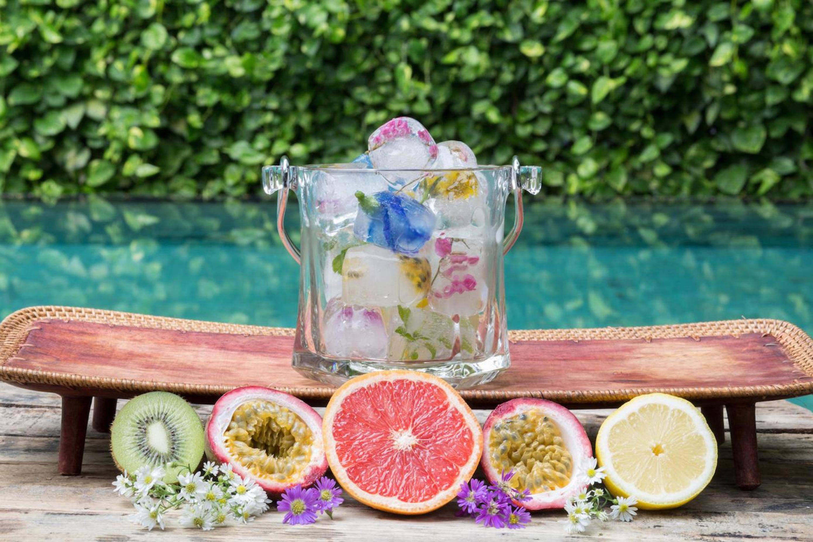 Flower-infused ice cubes and tropical fruit halves create an Instagram-worthy poolside moment, where vibrant passion fruit and ruby grapefruit meet artisanal refreshment under dappled sunlight.