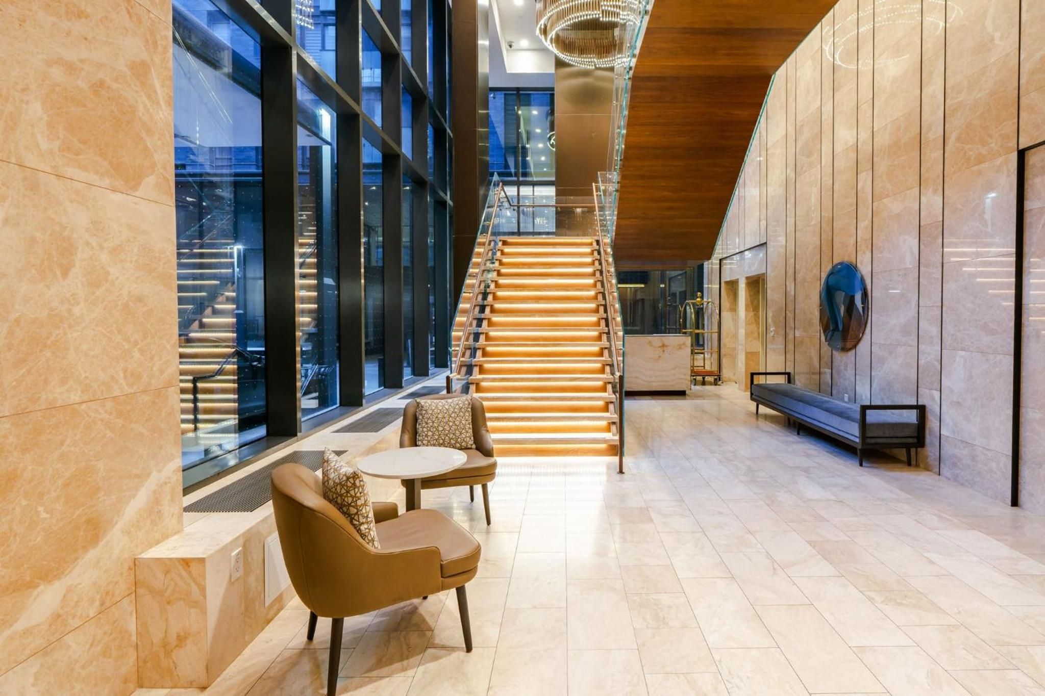 Warm travertine and illuminated wood stairs create an inviting lobby sanctuary where mid-century seating invites lingering beneath soaring glass walls.
