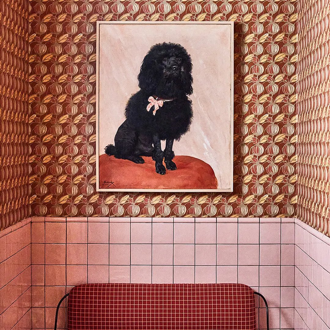 Whimsical wallpaper featuring repeating botanical motifs creates an unexpected gallery moment above the coral-tiled bathtub, where a vintage poodle portrait adds playful sophistication to this jewel-box bathroom.