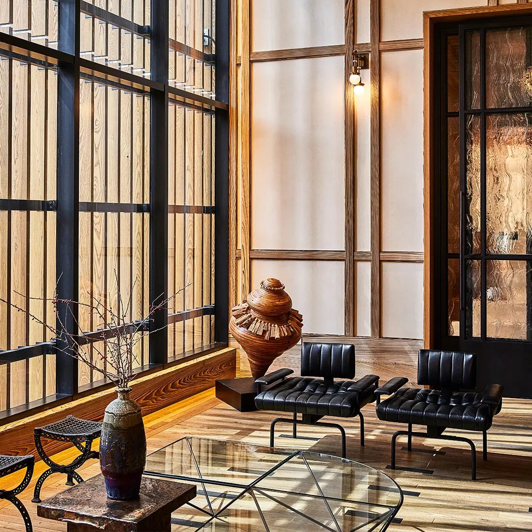 Sunlight streams through floor-to-ceiling windows, illuminating Barcelona chairs and artisanal pottery against warm wood paneling in this thoughtfully curated lobby sanctuary.