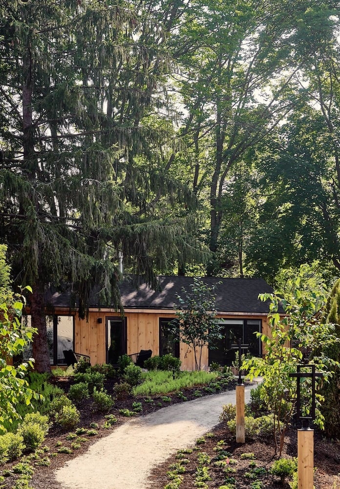 Natural cedar siding and thoughtfully landscaped gardens create an understated retreat beneath towering Maine pines, where gravel pathways lead to minimalist cabin-style accommodations.